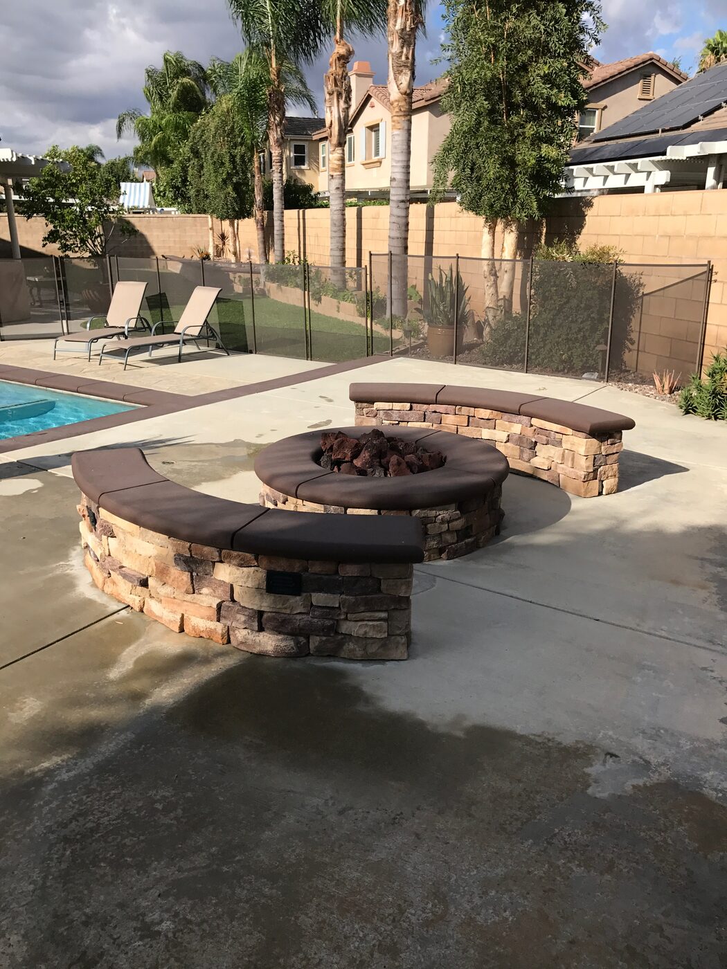 Round stone fire pit with curved cushioned seat walls beside a backyard pool